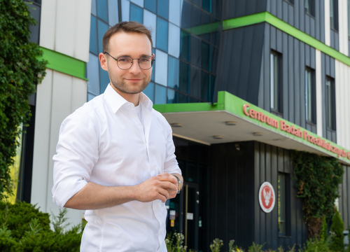 Młody mężczyzna, Karol Momot, pozuje do zdjęcia. W tle widać budynek z czarno-białą fasadą z elementami zieleni, czerwonym napisem: Centrum Badań Przedklinicznych CePT oraz godłem WUM.