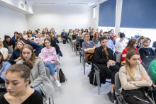 People sitting in a lecture theatre. A man in a suit stands in front of them. He is addressing a woman sitting to his left.