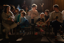 Grupa osób. Dzieci oraz dorośli. Siedzą na krzesłach w dużej sali. Światła w sali przyciemnione.