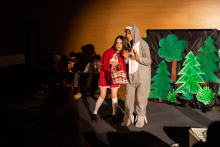 Two people in the foreground. The girl on the left wearing a red dress and red cape. The girl on the right wearing a bunny costume. Behind them a paper scenery showing a forest