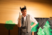 Young man. He has wolf ears on his head. He is dressed in a grey jacket and white shirt. He wears glasses.  In the background a paper scenery showing a forest