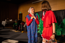 Two women. The woman on the left dressed in her grandmother's outfit: blue apron and red shirt. The woman on the right dressed in a red dress and red cape