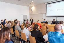 Grupa osób siedzących tyłem. Przed nimi stół konferencyjny. Siedzą przy nim kobieta i mężczyzna. Po prawej stronie stoi kobieta z mikrofonem w ręku. 