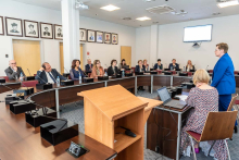 Several people are seated at horseshoe-shaped tables. In front of them is a rostrum with two women at it. One of them sits and the other stands and gives a lecture.