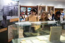 Na pierwszym planie gablota muzealna ze starodawnymi dokumentami. Za gablotą stoją dwie młode dziewczyny. W tle zdjęcia medyczne i historyczne.