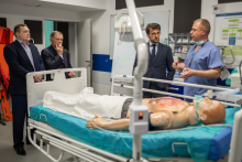 Four men, one wearing a blue medical gown. They are standing around a hospital bed on which a phantom of a patient lies.