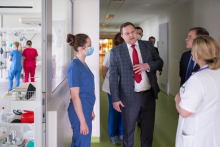 Grupa osób na szpitalnym korytarzu - lekarka w białym fartuchu odwrócona tyłem, młoda kobieta w niebeskim stroju medycznym, i dwóch mężczyzn w garniturach.