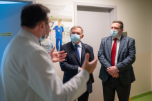 A doctor in a white apron is explaining something to two men in suits and with surgical masks on their faces.