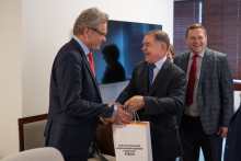 Three men in suits. The two in the foreground shake hands and hand over a bag.