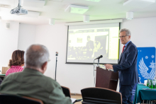 Mężczyzna w garniturze - rektor, stoi za mównicą i przemawia. Za nim, z boku, ekran z prezentacją oraz rollup.