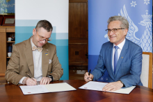 Two men sit at a table. They sign documents.