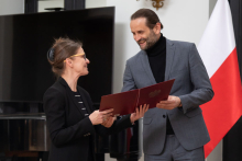 Mężczyzna w szarym garniturze i kobieta w czarnym garniturze, razem trzymają dyplom. 