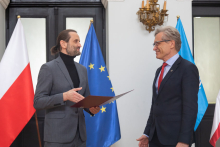 Dwaj mężczyźni stoją naprzeciwko siebie , jeden trzyma w ręku dyplom. W tle flaga Polski i Unii Europejskiej.