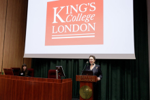 Kobieta stojąca za mównicą. Ubrana oficjalnie. Za nią ekran, a na ekranie logotyp - czerwony kwadrat z białymi literami - napis King's College London