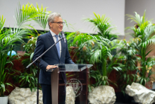 An elderly man at the lectern. Green palm trees in pots in the background.