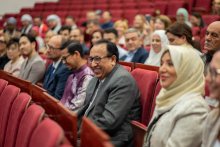 Audience. Many people are seated in red chairs. In the foreground a woman in a hijab, next to her men in suits.