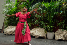 A dark-skinned woman in a raspberry-colored dress and a turban on her head sings and dances with her arms outstretched. 