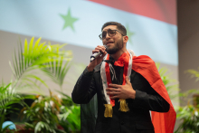 A young boy wearing glasses with a Syrian flag on his shoulders sings into a microphone.