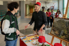 A boy wearing a traditional red Turkish cap at a booth selling leaflets and various items. On the other side, a student approaches the booth.