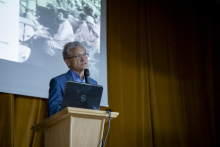 An elderly man at the lectern, speaks into a microphone. Behind him you can see a section of a screen with black-and-white photos.