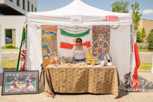 A girl wearing a white blouse with buff sleeves, jeans and dark glasses is standing at a booth with a sign reading Iran.