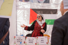 A young girl with an "arafat" on her head and traditional attire. She makes an inviting gesture to the booth. Behind her is the flag of Palestine.