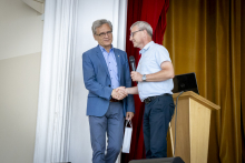 Two men in front of a lectern in a lecture hall. They shake hands.