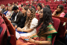 Audience. Many people are seated in red chairs. In the foreground a woman in a hijab, next to her men in suits.