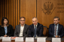 Stół na sali konferencyjnej. Siedzą przy nim prelegenci. Widać kobietę i trzech mężczyzn.