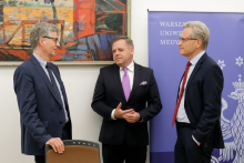 Three men in suits in an American set. They are talking. In the background on the poster is the logo of Warsaw Medical University.