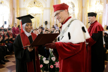In the foreground, a man in a red toga with a white collar presents a diploma to a girl dressed in a black toga and biretta.