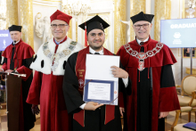 Three men smile for a photograph. The man in the middle, wearing a black toga and biretta holds a diploma. 