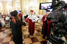 In the foreground, to the right, a standing camera with recording on. In front of the camera, a man in a red toga with a white collar shakes hands with a young woman dressed in a black toga and biretta.