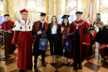 Group of six standing. In the background a man standing behind a lectern wearing a black toga. In the foreground 3 men and two women. They are smiling for the photograph. Two people are holding navy blue gift bags.