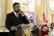 A man in a black suit and red tie. He stands behind the lectern. With one hand he holds the microphone and speaks. Behind him, the WUM banner can be seen in the background.