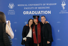 Three people, a young woman wearing a toga and biretta, and a man and a woman pose for a photo against a blue background with the WUM logo and information about the Diplomatatorium.