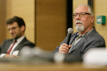 Two men sit at a conference table. In the foreground an older man with a beard and moustache is dressed in a grey suit. In the background is the blurred figure of the other seated man.