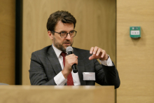 Close-up of a man with dark hair wearing glasses speaking into a microphone. He gestures with his left hand.