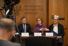  Conference table. Two men are sitting and a woman is gesturing into a microphone.