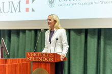 A woman, blonde, wearing a white jacket stands behind the lectern and speaks. Behind her a screen, on the screen - the KRAUM logo.