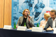 Young man with glasses. He is seated behind a table. He holds a microphone in his left hand and speaks. Next to him sits a woman with fair hair and a middle-aged man. Behind them a rollup.