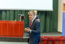 A man, slim, dressed in a jacket. He stands in the middle of the room and speaks to the audience. Behind him is a lectern and a green curtain.