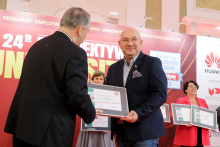 In the foreground, two men. One, with his back turned to the camera, hands the other - facing forward, wearing glasses and a suit - a diploma.