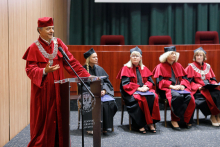 Uroczystość wręczenia dyplomów ukończenia studiów. Na pierwszym planie mężczyzna (prorektor) w czerwonej todze przy mównicy. Z tyłu siedzą cztery osoby również w togach.
