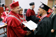 Mężczyzna (prorektor) w czerwonej todze wręcza absolwentom dyplomy.