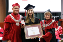 Graduation ceremony. Three people can be seen. In the center a female graduate with a framed diploma. On the right a man (pro-rector) on the left a woman (dean). Both wearing red togas.