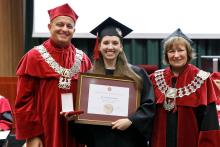 Graduation ceremony. Three people can be seen. In the center a female graduate with a framed diploma. On the right a man (pro-rector) on the left a woman (dean). Both wearing red togas.