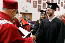 A man (pro-rector) in a red toga presents diplomas to graduates.
