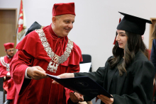 A man (pro-rector) in a red toga presents diplomas to graduates.