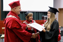 A man (pro-rector) in a red toga presents diplomas to graduates.